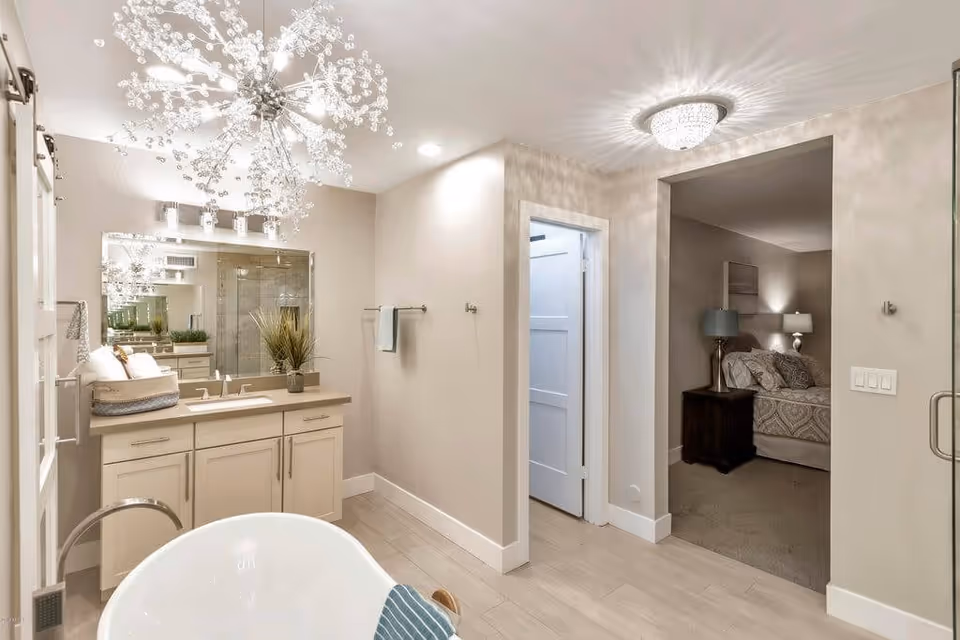 A modern bathroom with a white freestanding bathtub in the foreground, a vanity with a large mirror and sink, and a glass-enclosed shower. The bathroom has light-colored walls and flooring, with a decorative chandelier on the ceiling. An open doorway leads to a bedroom with a bed, nightstands, and lamps.
