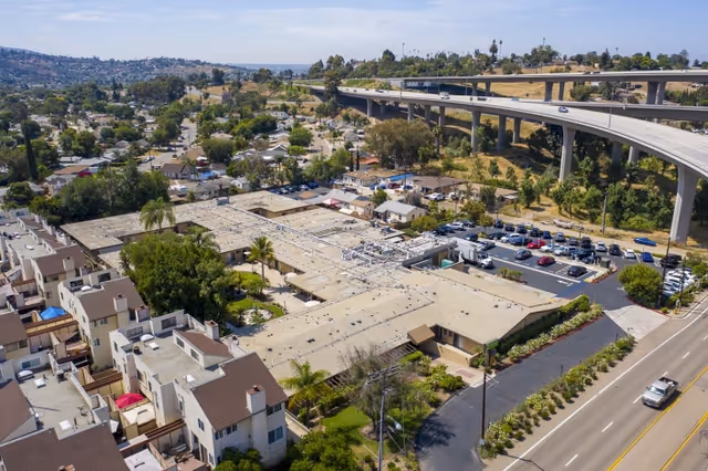 Aerial view of Lemon Grove Care And Rehabilitation Center showing a large single-story building with a flat roof, surrounded by parking lots, trees, and nearby residential houses. A highway overpass is visible in the background along with a hilly landscape under a partly cloudy sky.
