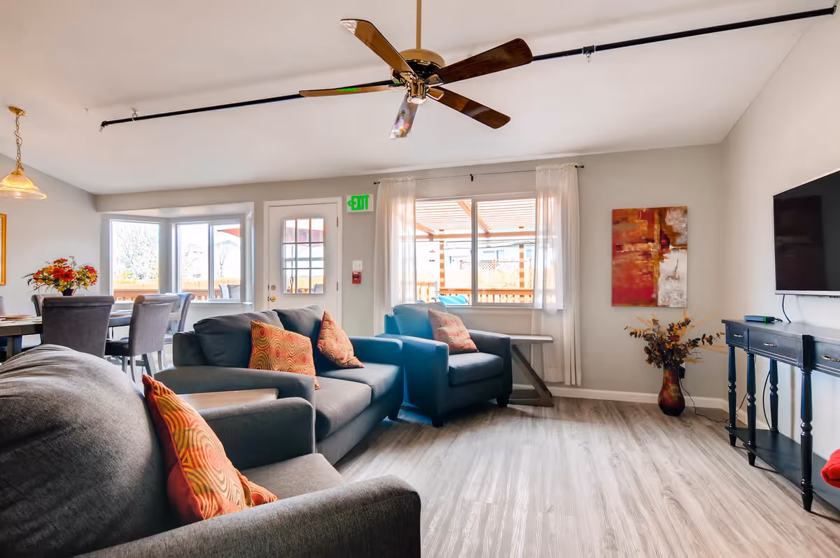 A bright and spacious living room in an assisted living facility featuring a ceiling fan, multiple armchairs and sofas with patterned cushions, a flat-screen TV mounted on the wall, a console table, a dining area with a table and chairs, large windows with sheer white curtains, and a decorative vase with flowers.