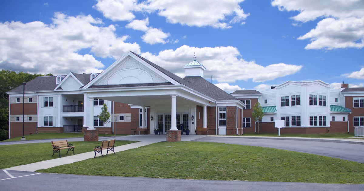 Exterior view of the Hannah B.G. Shaw Home building under a partly cloudy blue sky, featuring a covered entrance with white columns, brick and white siding facade, a driveway, green lawn, and two benches near the walkway.