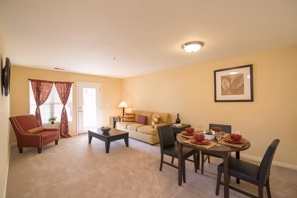 A cozy living room and dining area in Marwood Senior Apartments featuring a beige sofa with colorful cushions, a red armchair, a dark wooden coffee table, and a round dining table set with four place settings including red bowls and glasses. The room has beige walls, carpeted floor, a window with red patterned curtains, a door with glass panels, a table lamp, and a framed leaf artwork on the wall.