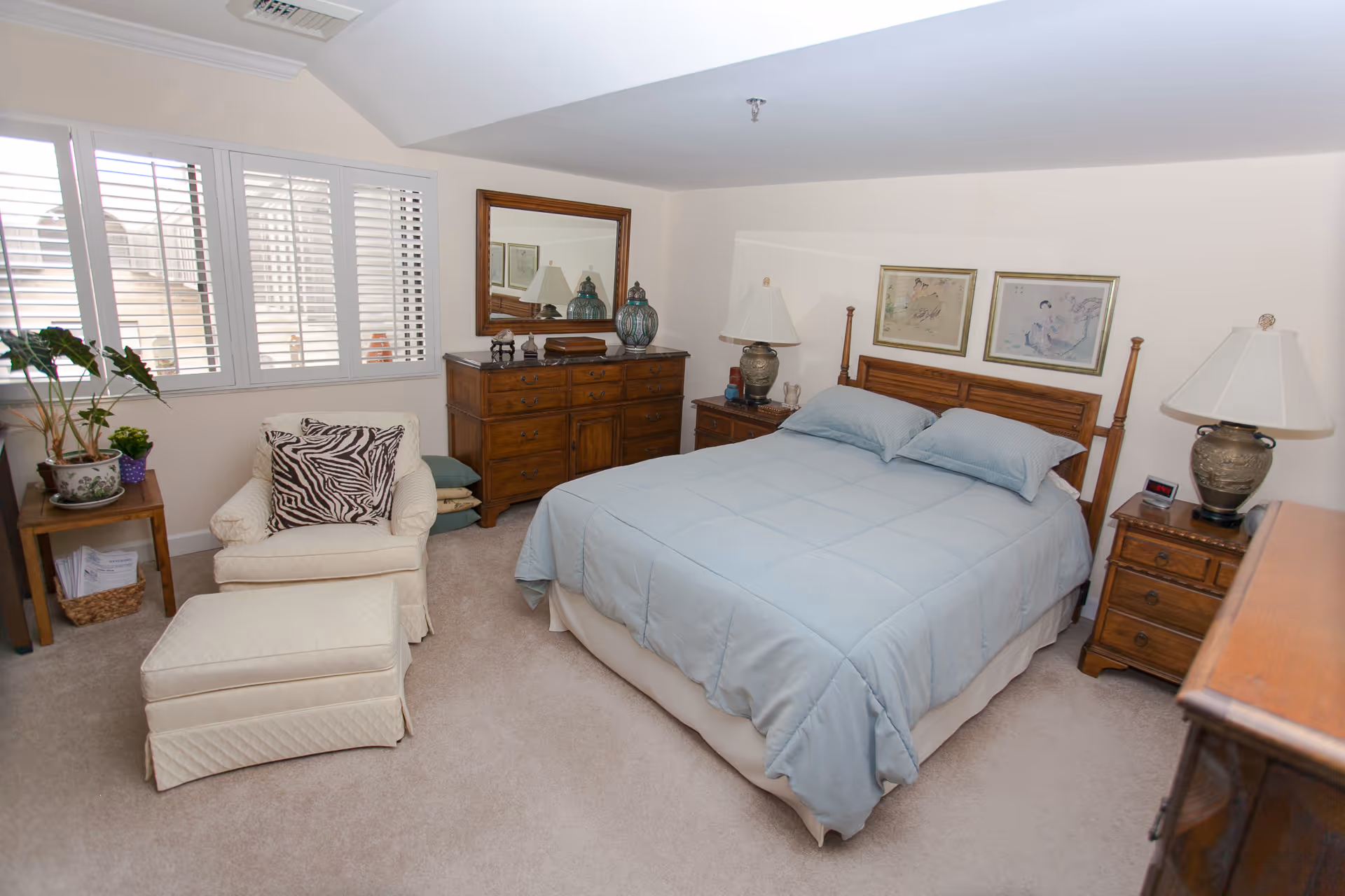 Bright bedroom with a wooden bed dressed in light blue bedding, matching nightstands and dresser, and a cushioned armchair by shuttered windows.