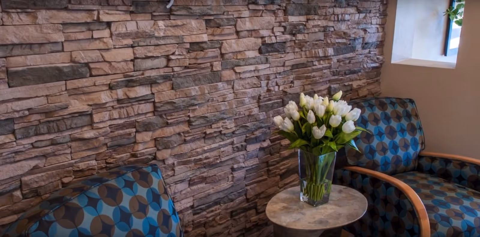 Two patterned armchairs with wooden armrests positioned around a small round table holding a clear vase filled with white tulips, set against a textured stone wall with a small window on the right.
