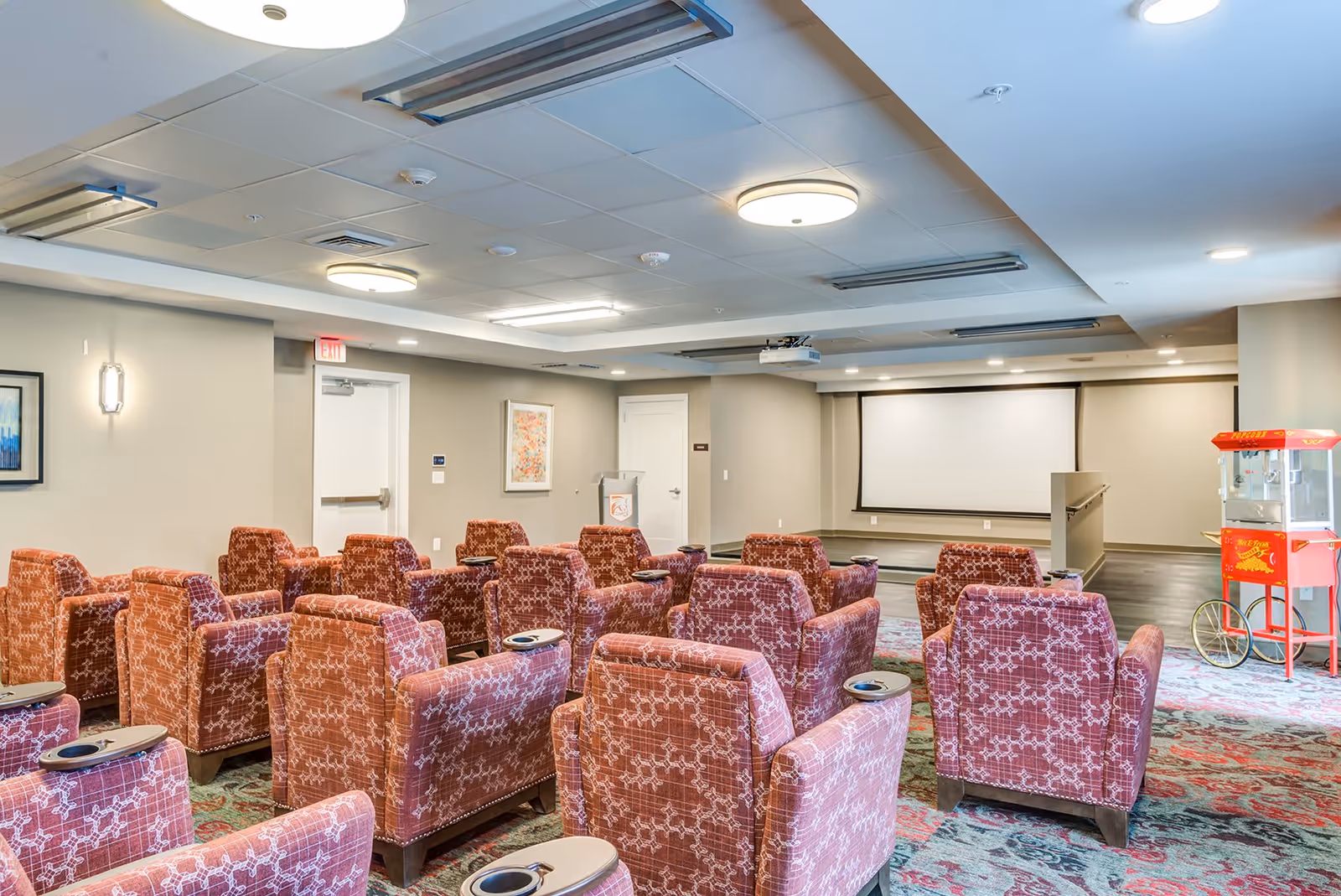 A small theater room with multiple rows of red patterned armchairs facing a large white projection screen. The room has a carpeted floor with a colorful design, neutral-colored walls with framed artwork, ceiling lights, and a red popcorn machine on the right side near the screen.