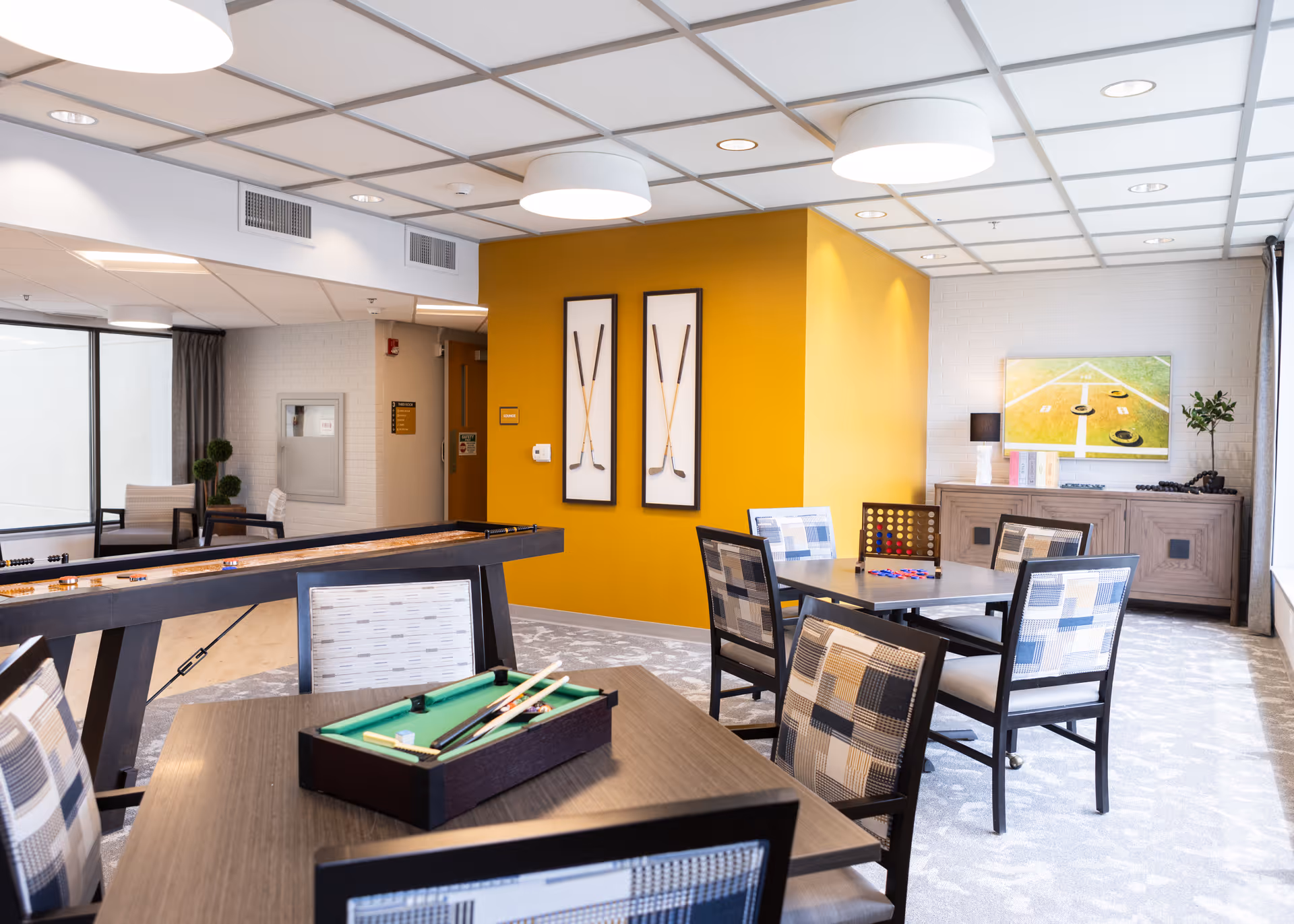 A bright and modern common area in a senior living facility featuring several tables and chairs with patterned upholstery. On one table, there is a small pool table game set. In the background, there is a shuffleboard table and a yellow accent wall with two framed pictures of crossed golf clubs. A cabinet with a lamp, books, and a plant is against the far wall, which also has a TV displaying a shuffleboard game. Large windows allow natural light to fill the room.