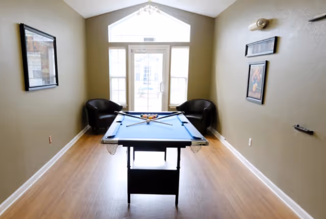 A small game room with a pool table centered in the room, two black armchairs positioned near a glass door with windows on either side, light wood flooring, beige walls with framed pictures, and a high triangular window above the door letting in natural light.