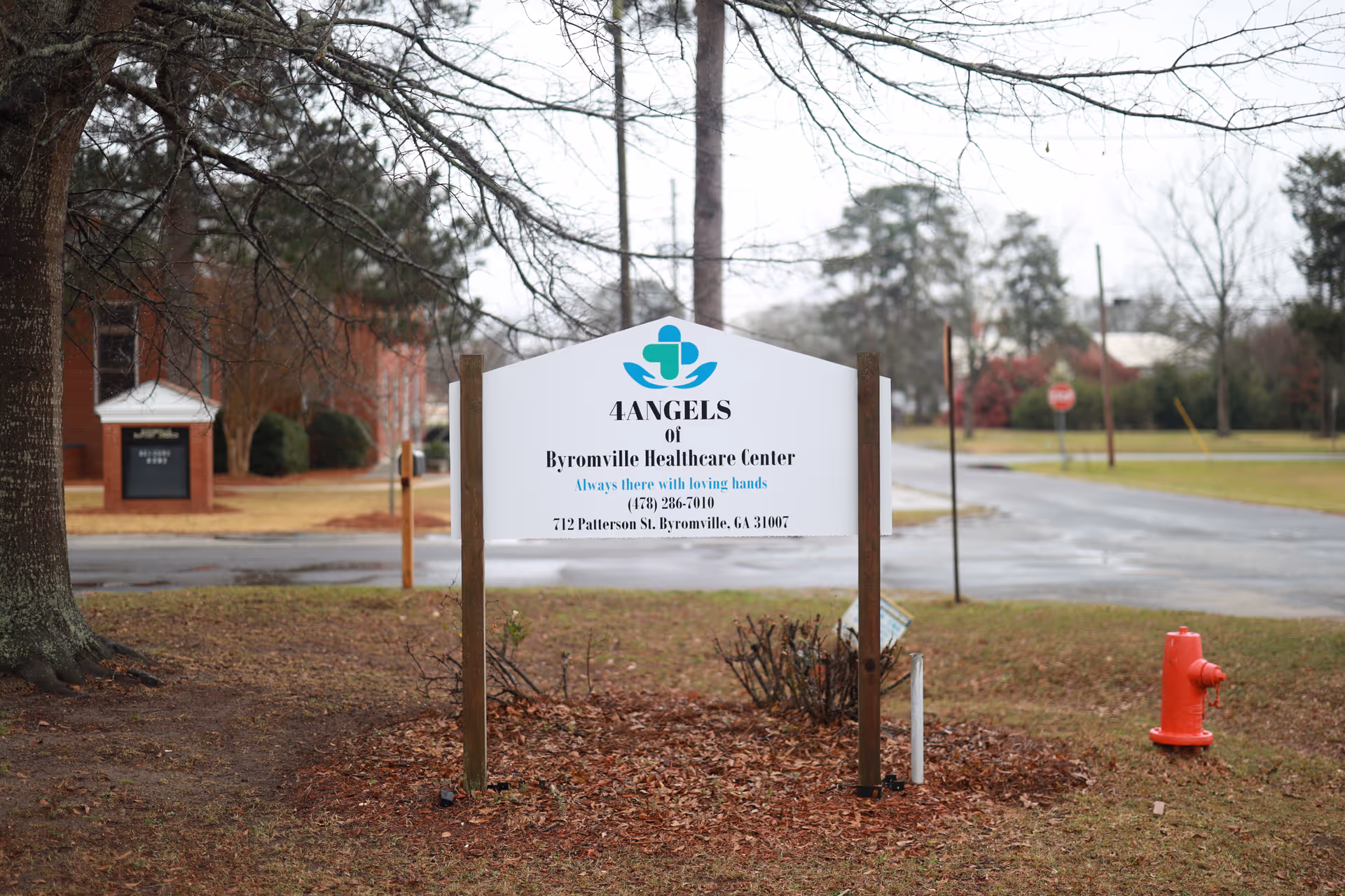 Outdoor view of a white sign for 4ANGELS of Byromville Healthcare Center with contact information, set on a grassy area with trees and a red fire hydrant nearby. A road and some buildings are visible in the background.