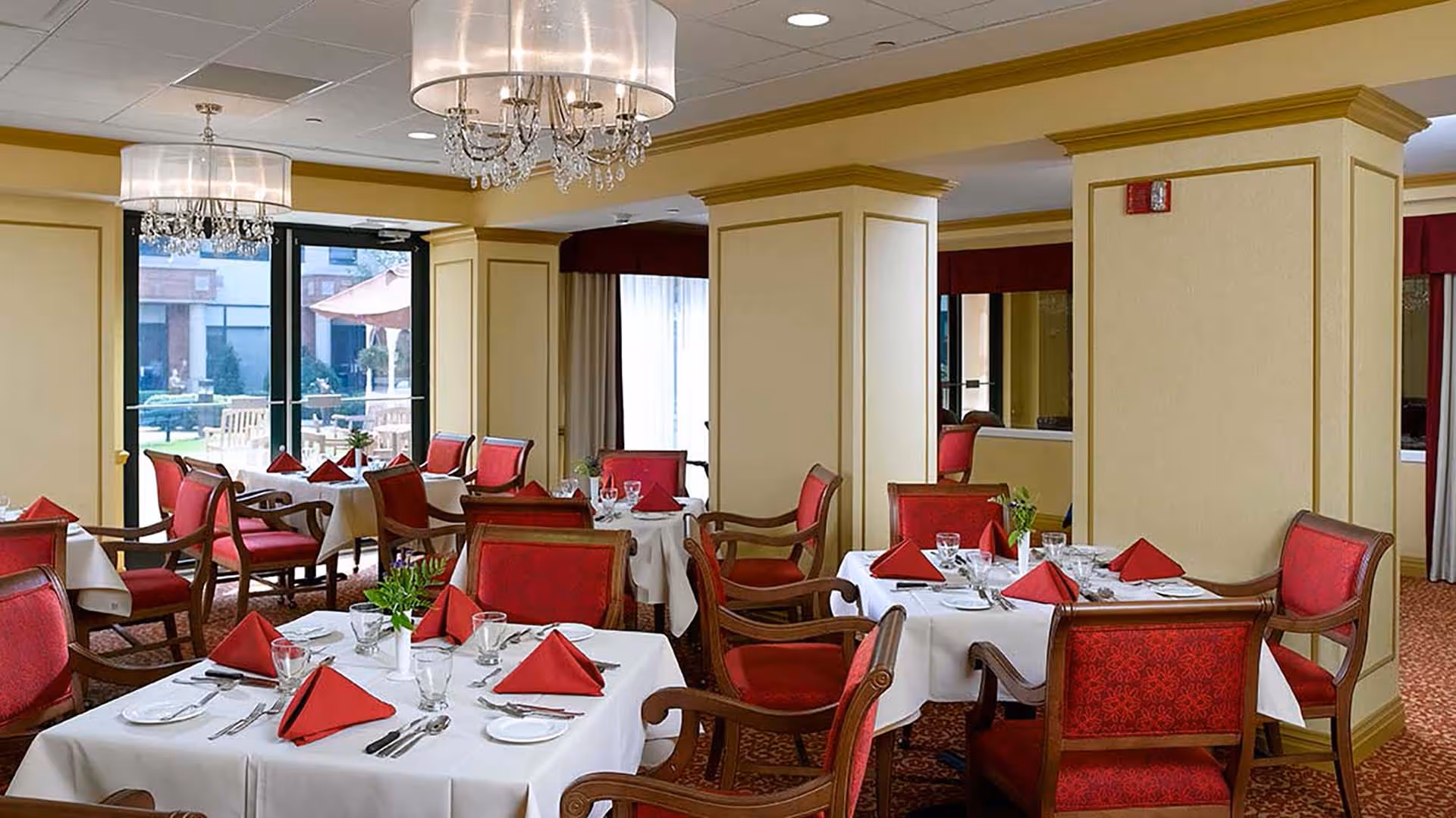 Elegant dining room with tables covered in white tablecloths, set with red folded napkins, glassware, and silverware. The room features red upholstered wooden chairs, beige walls with gold trim, chandeliers with crystal accents, and large windows showing an outdoor patio area with umbrellas and seating.