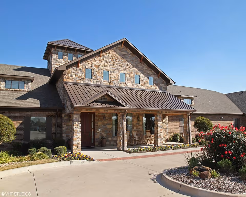 Exterior view of a stone and brick building with a peaked roof and a covered entrance supported by stone columns. There are rocking chairs on the porch and landscaped bushes and flowers around the driveway under a clear blue sky.