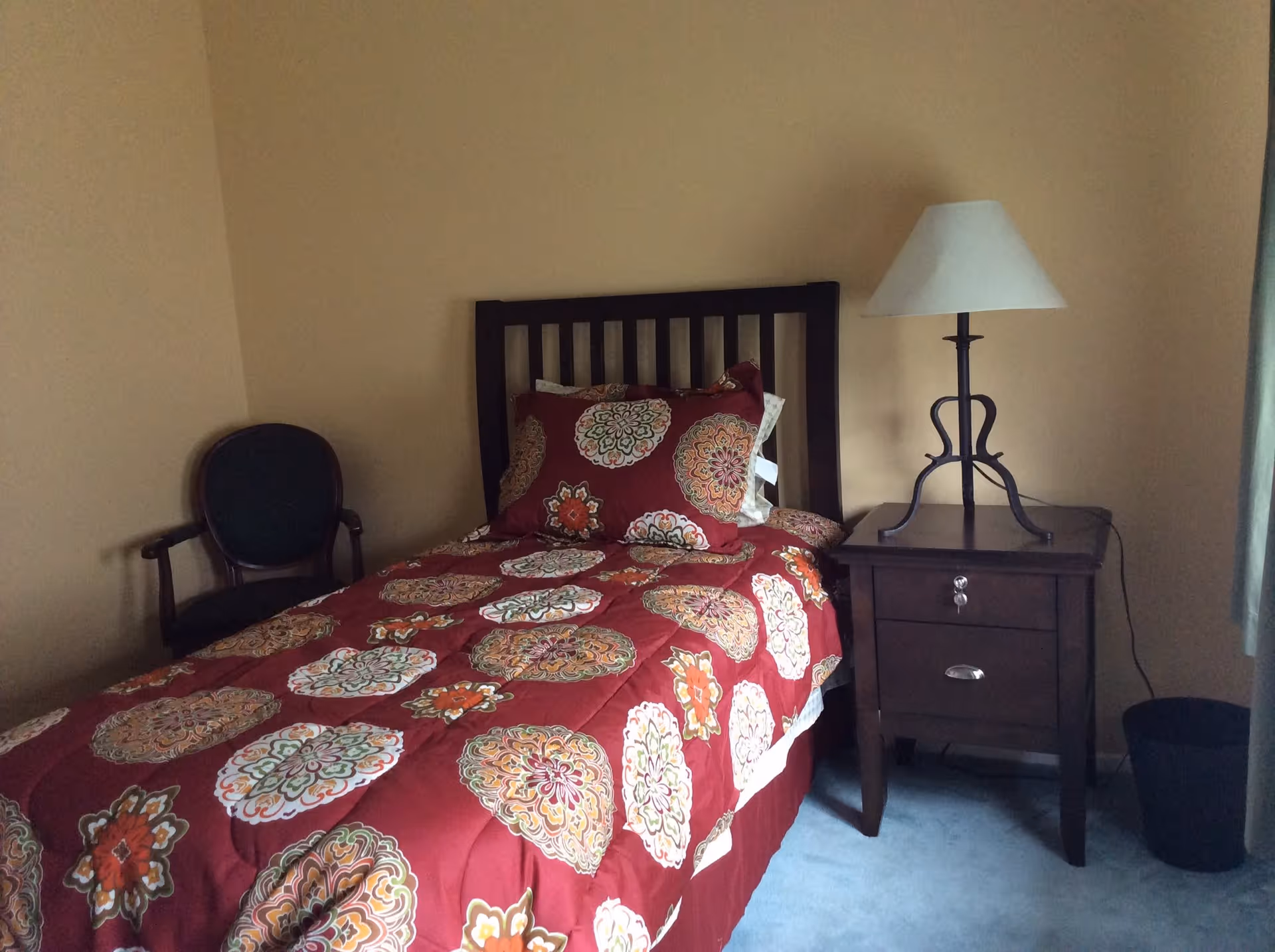 A bedroom with a single bed featuring a red comforter with colorful circular patterns. Next to the bed is a dark wooden nightstand with two drawers and a lamp with a white lampshade. There is a dark wooden chair with a green cushion in the corner, and a window with light curtains on the right side.