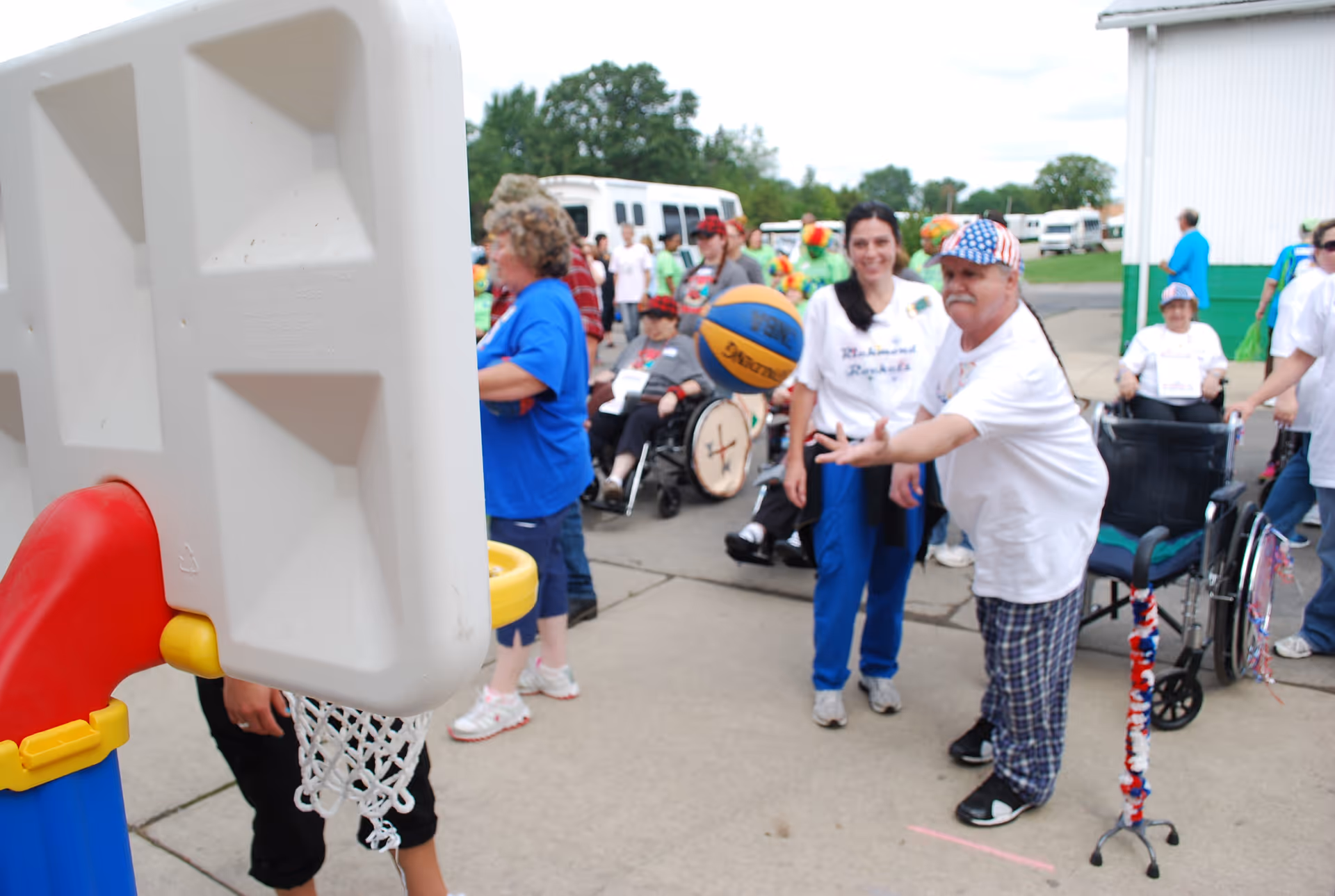A group of people, including elderly individuals in wheelchairs and caregivers, are outdoors playing basketball with a small plastic basketball hoop. One man is actively throwing the basketball while others watch and smile. The setting appears to be a paved outdoor area near a building with green and white walls.