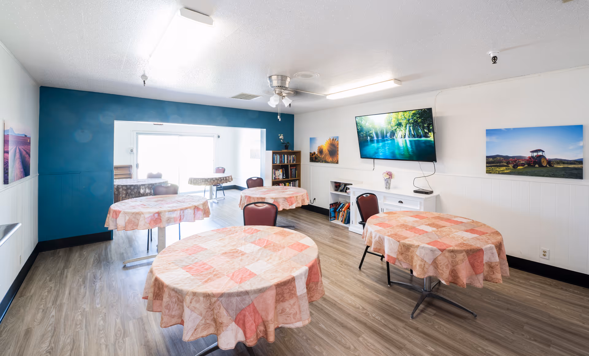 A bright room with four round tables covered with pink and beige checkered tablecloths, each surrounded by chairs. The room has wood flooring, white walls with a teal accent wall, and a ceiling fan with lights. A large flat-screen TV is mounted on the wall displaying a nature scene. There are framed pictures on the walls and a bookshelf with books and decorative items.