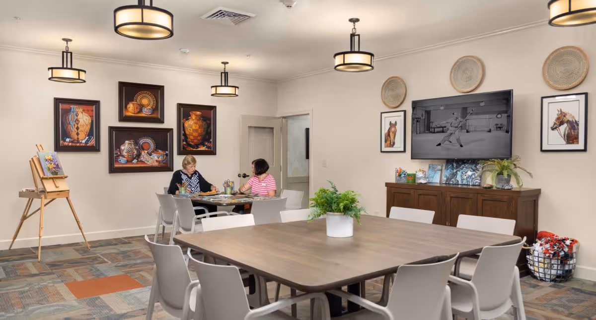 Communal activity/dining room with a large table and chairs, framed artwork and a wall-mounted TV, an easel, and two people doing crafts at a side table.