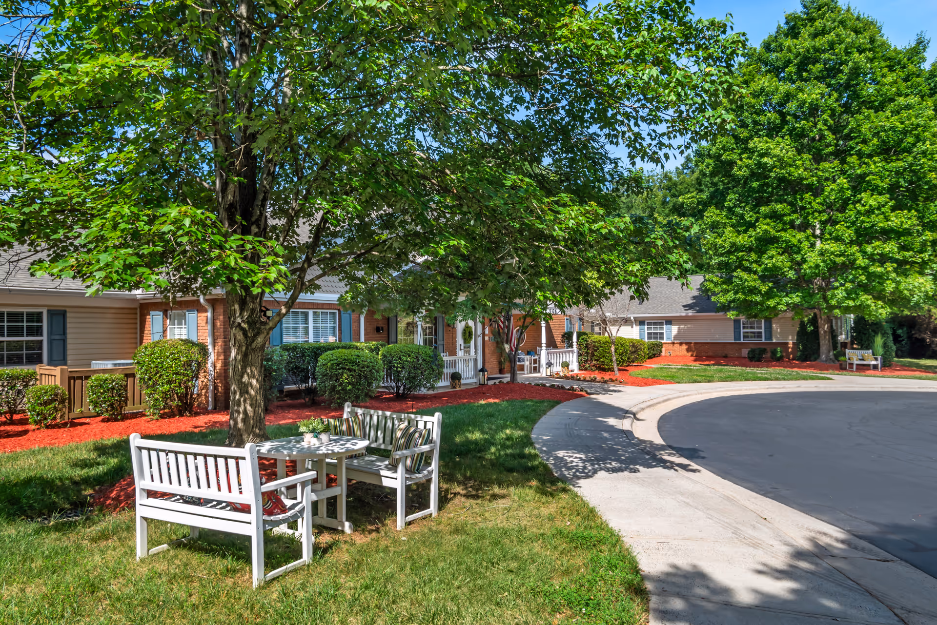 Front exterior of a single-story senior living building with landscaped lawn, trees, white outdoor benches around a table and a curved driveway.
