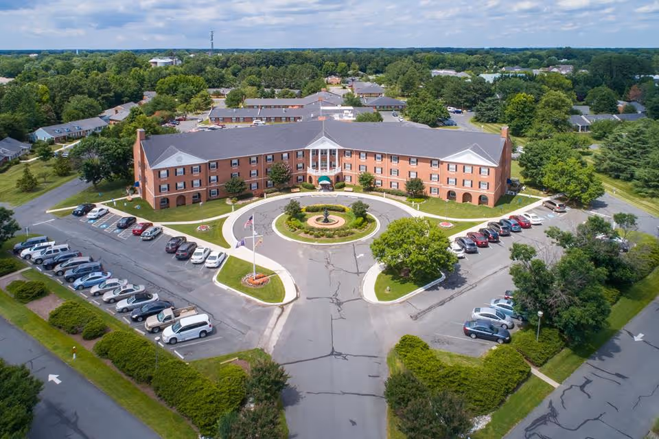 Aerial view of a large, three-story brick senior living facility with a circular driveway and a landscaped roundabout featuring a fountain in front. The building is surrounded by parking lots with numerous cars and green trees and lawns.