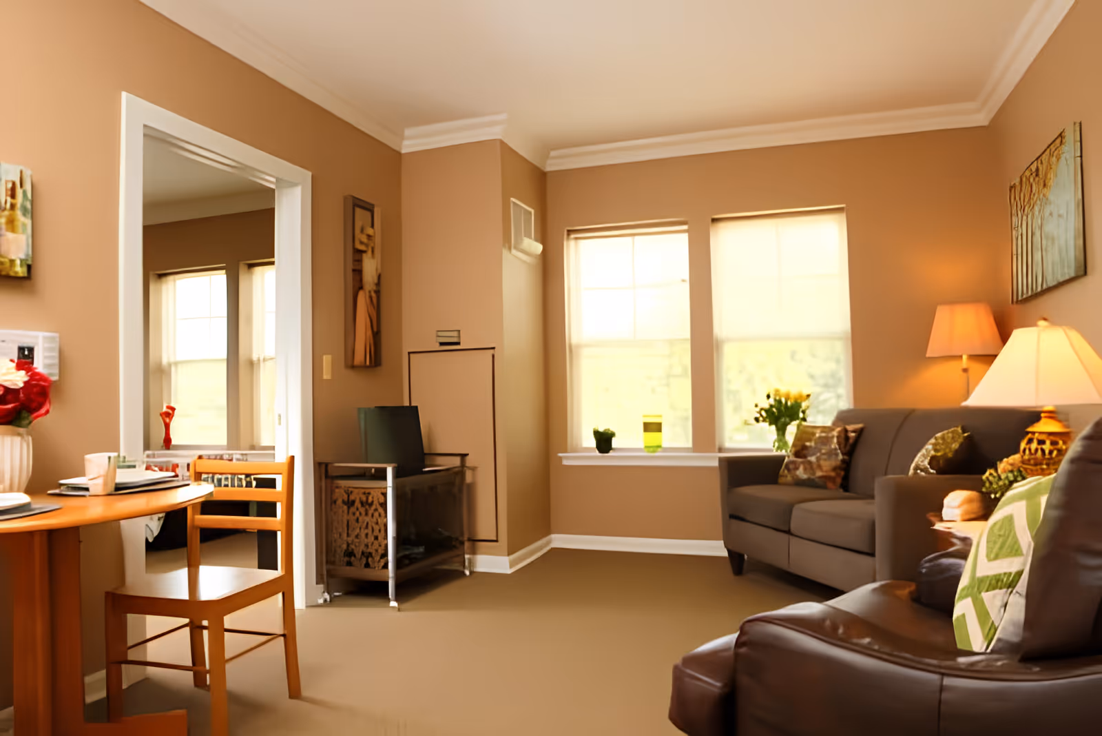 A cozy living room with beige walls and carpet, featuring a brown sofa with decorative pillows, a leather armchair with a green patterned pillow, a wooden dining table with chairs, two large windows with blinds, a side table with a lamp, and wall art. The room is warmly lit and has a doorway leading to another room.