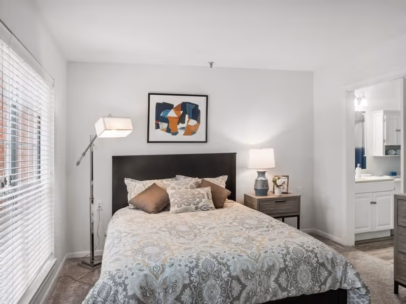 A bedroom with a bed featuring a patterned bedspread and several pillows. There is a floor lamp to the left of the bed and a nightstand with a table lamp and a small plant to the right. A framed abstract artwork hangs above the bed. To the right, an open doorway leads to a bathroom with white cabinetry and a mirror.
