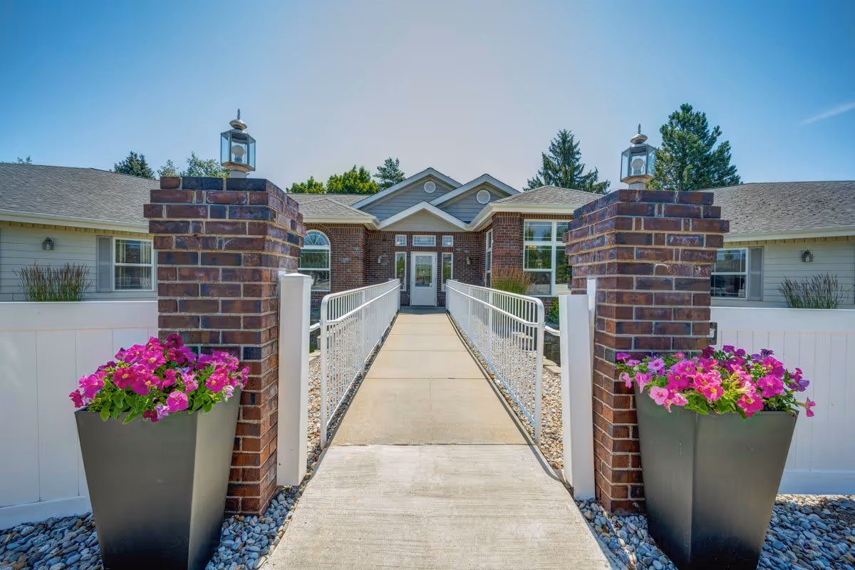 Front entrance of a brick and siding building with a concrete walkway flanked by two brick pillars topped with lanterns and large planters filled with pink and purple flowers under a clear blue sky.