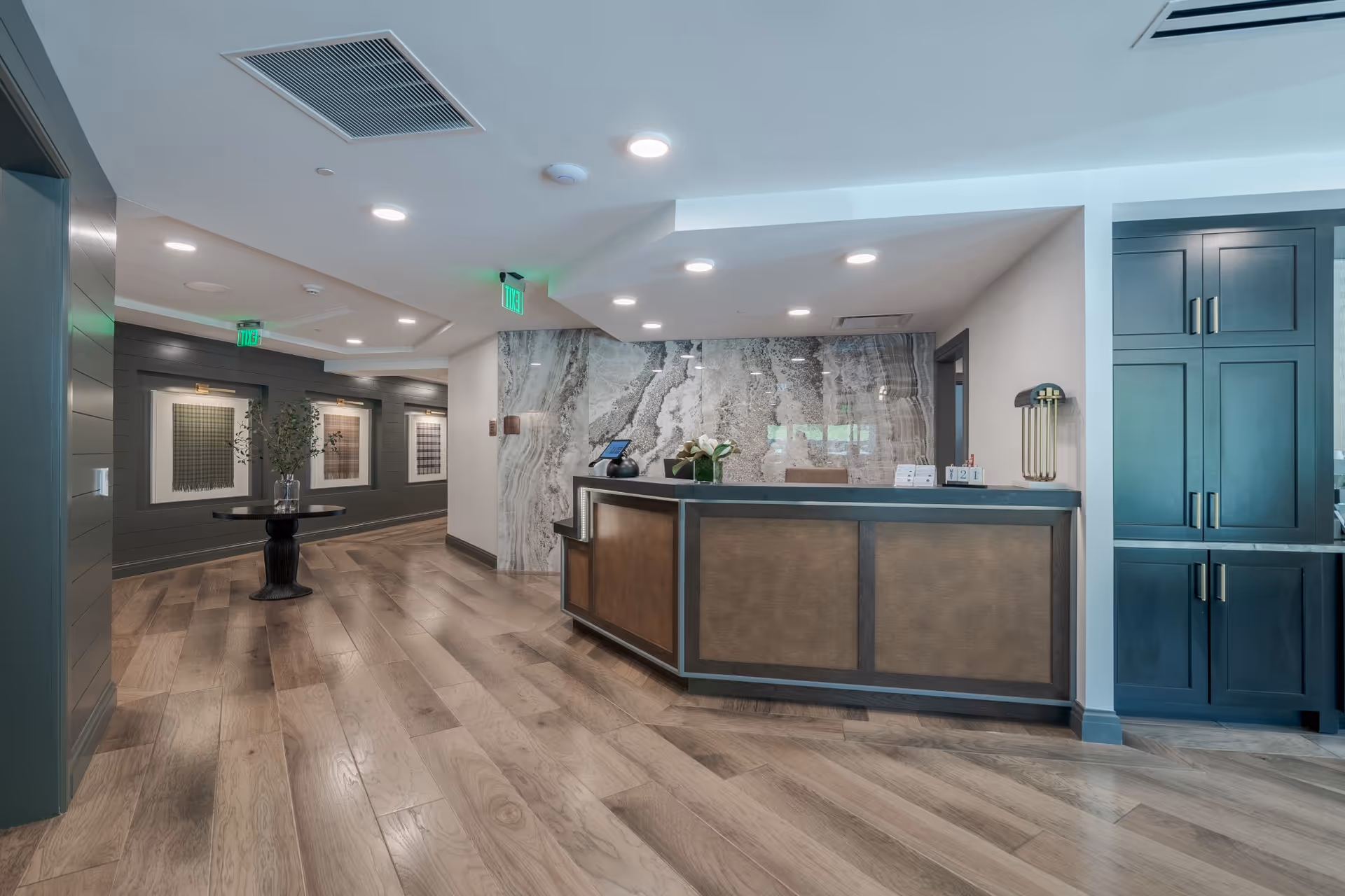 A modern reception area with a wooden floor, a front desk with a marble backdrop, and a hallway with framed artwork on the walls. The space is well-lit with recessed ceiling lights and has a table with a vase of flowers in the hallway.