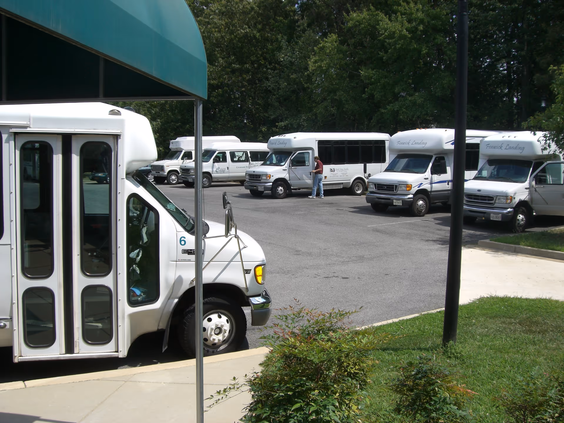 Parking lot with several white shuttle vans, some labeled Fenwick Landing, parked near a green awning and surrounded by trees and grass. A person is standing near one of the vans.