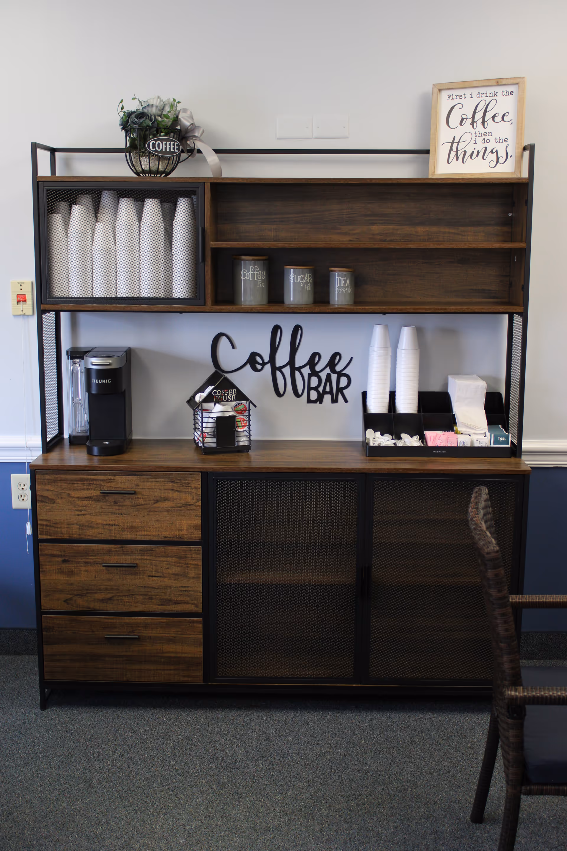 A coffee bar setup with a wooden and metal shelving unit. The top shelf holds a small plant with a 'COFFEE' sign and a framed sign that reads 'First I drink the Coffee, then I do the things.' The middle shelf has containers labeled Coffee, Sugar, and Tea. The bottom shelf has a Keurig coffee maker, a small decorative coffee house, stacks of white disposable cups, and compartments with coffee creamers, sugar packets, and napkins. The wall behind the unit has the words 'Coffee Bar' in black decorative lettering. A wicker chair is partially visible on the right side.