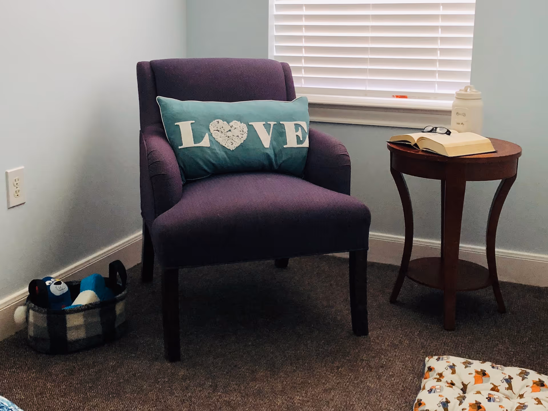 Purple upholstered chair with a teal 'LOVE' pillow beside a small wooden side table holding an open book and glasses in a softly lit room.