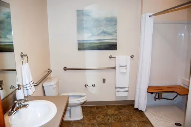 A clean and accessible bathroom with a sink and countertop on the left, a toilet in the center, and a walk-in shower with a wooden bench and white curtain on the right. There are grab bars near the toilet and shower, a towel rack with a white towel, and a landscape painting on the wall above the toilet.