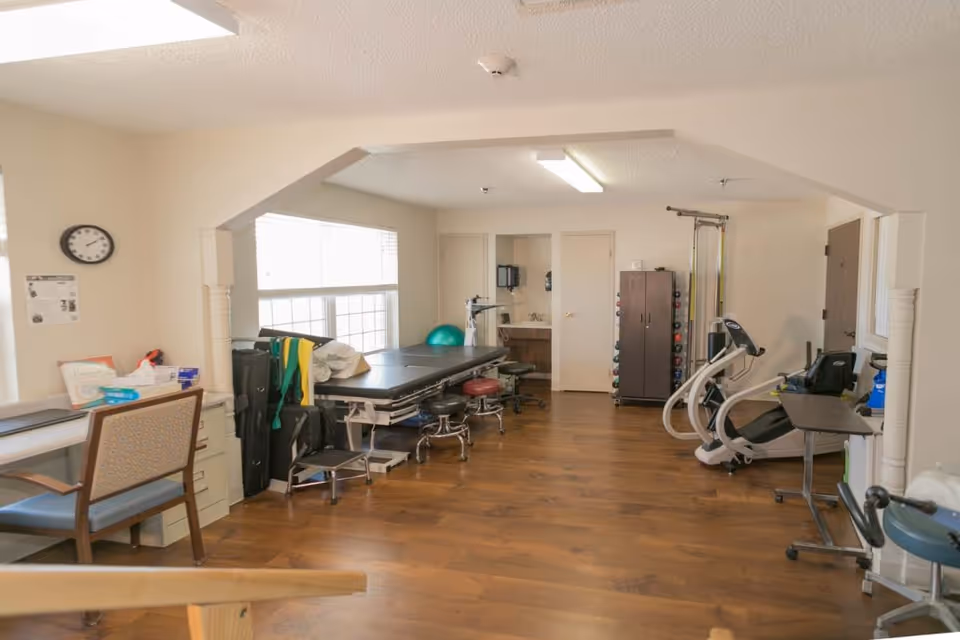 Bright therapy room with treatment tables, exercise machines, free weights and chairs on wood flooring.