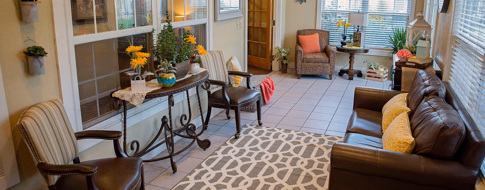 A cozy living room area with a brown leather sofa adorned with yellow pillows, two striped armchairs, and a patterned armchair near a window. A decorative console table with potted plants and flowers sits against the wall with large windows. The floor is tiled with a geometric patterned rug in the center, and the room is well-lit with natural light coming through the windows.