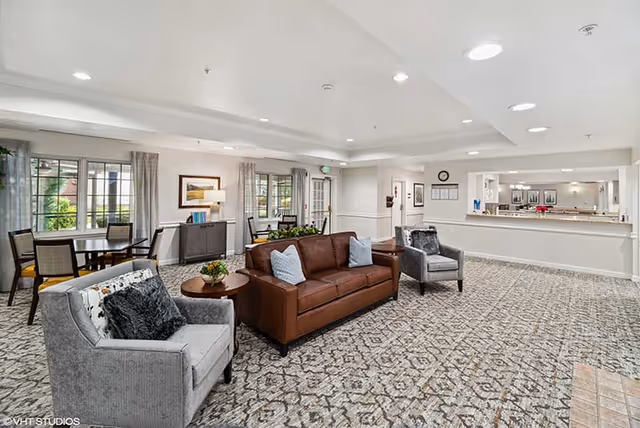 Bright communal living area with a leather sofa, armchairs, dining table, and a reception counter in a senior living facility.