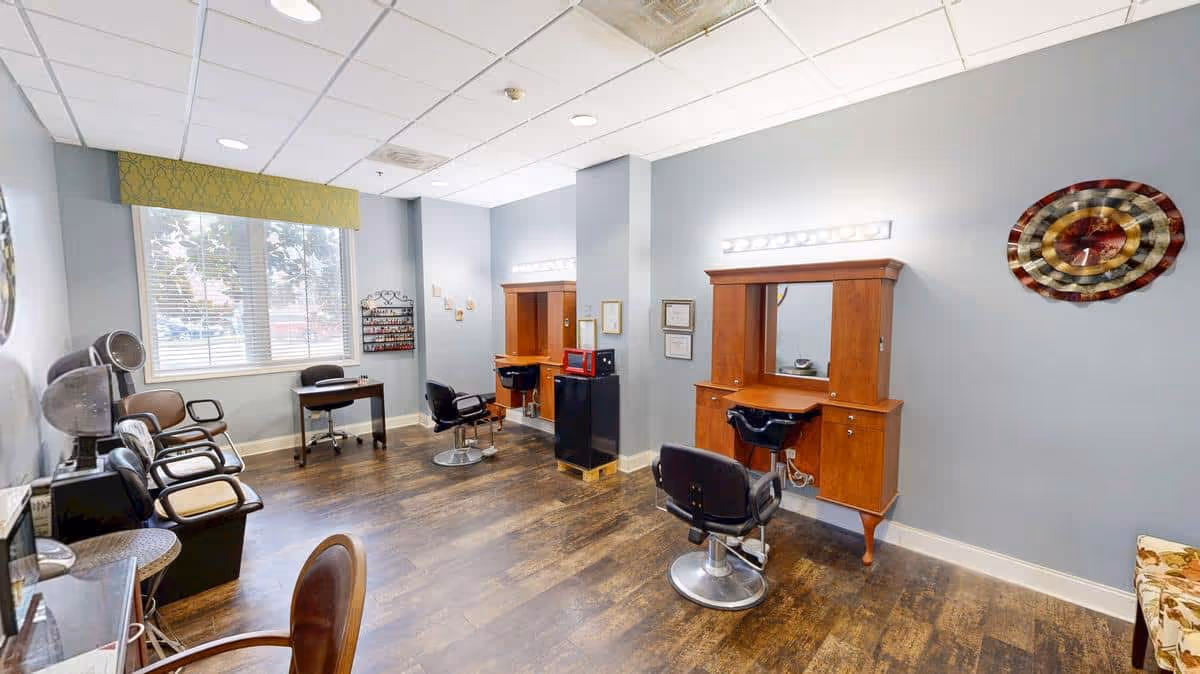 Interior view of a salon area with two wooden styling stations, each with a mirror and a black salon chair. There are additional chairs along the left wall, a small desk near a window with blinds, and a decorative wall clock on the right wall. The floor is wooden and the walls are painted light blue.