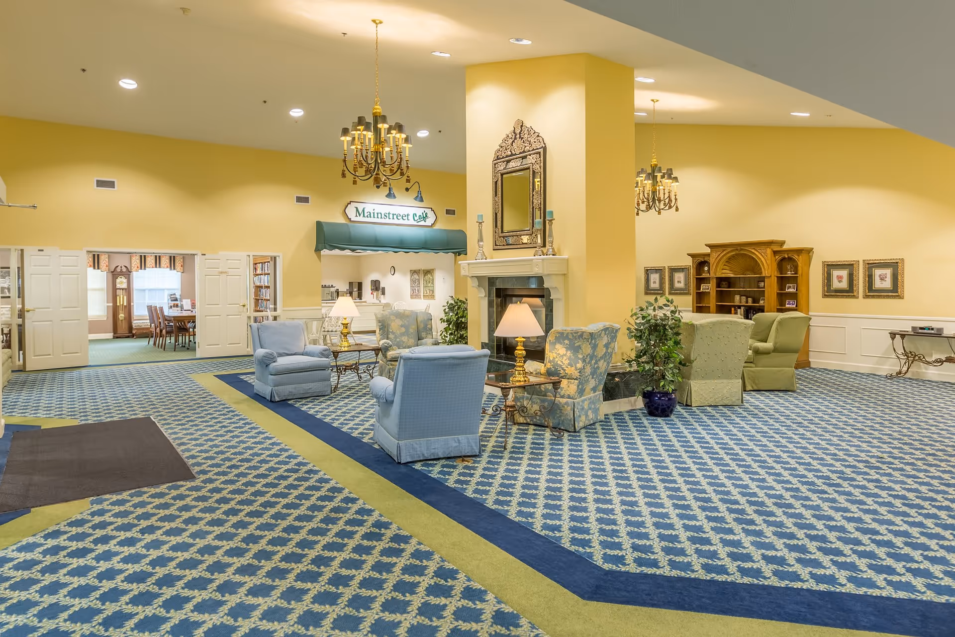 Spacious and well-lit common area in a senior living facility featuring patterned blue and green carpet, multiple armchairs arranged around a central fireplace with a decorative mirror above it, two chandeliers hanging from the ceiling, a small table with a lamp, potted plants, and a sign reading 'Mainstreet Cafe' above a counter area. In the background, there is an open doorway leading to a dining room with tables and chairs.