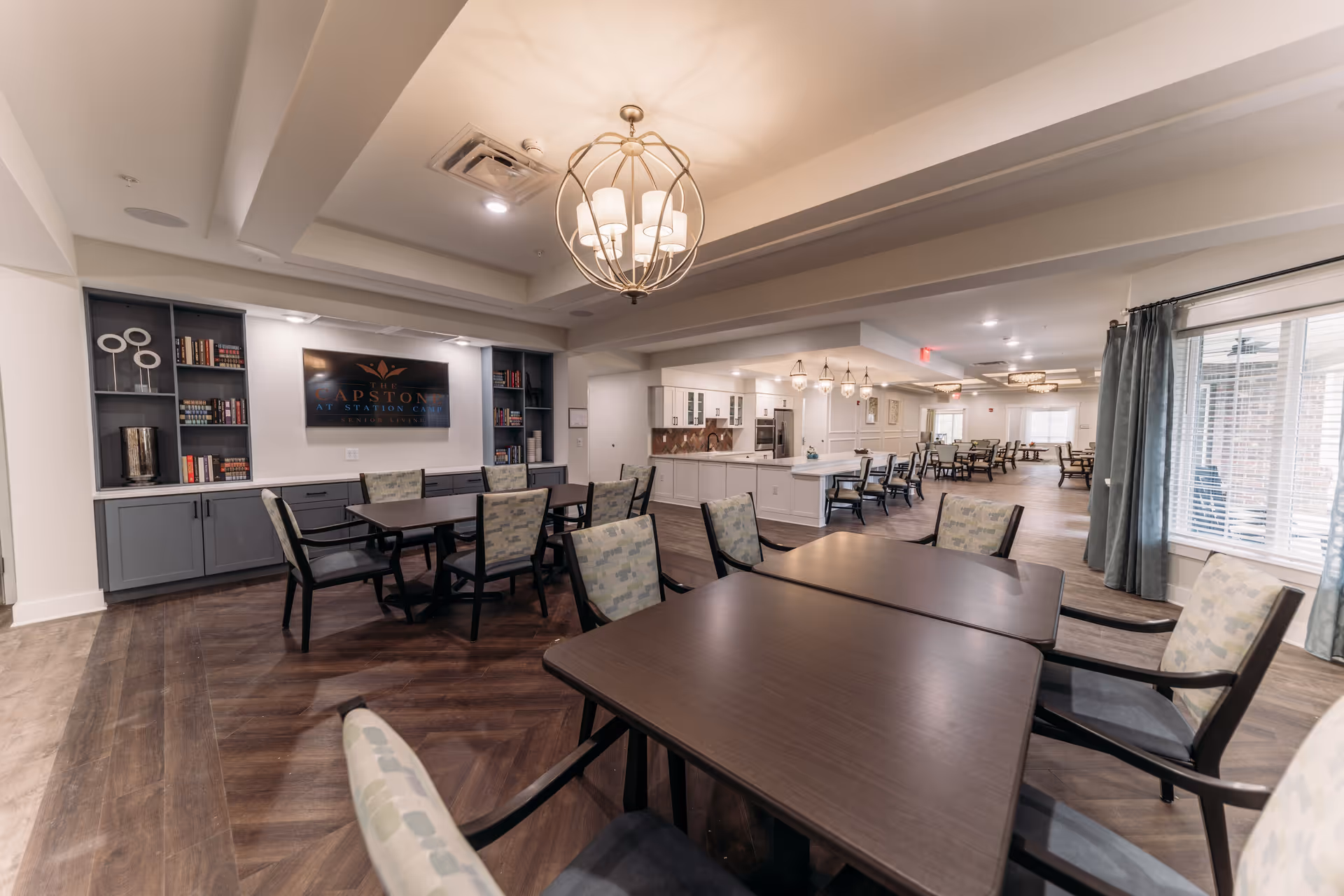 Spacious communal dining and activity room with multiple tables and chairs, built-in shelving and a kitchenette, featuring a sign that reads "Capstone at Station Camp".