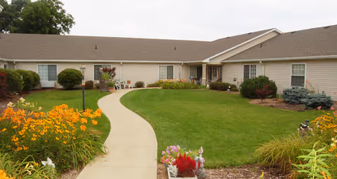 A single-story building with beige siding and a brown roof surrounding a green lawn with a curved concrete walkway leading to the entrance. The lawn is bordered by colorful flower beds with yellow, red, and purple flowers, and there are shrubs and small trees near the building.