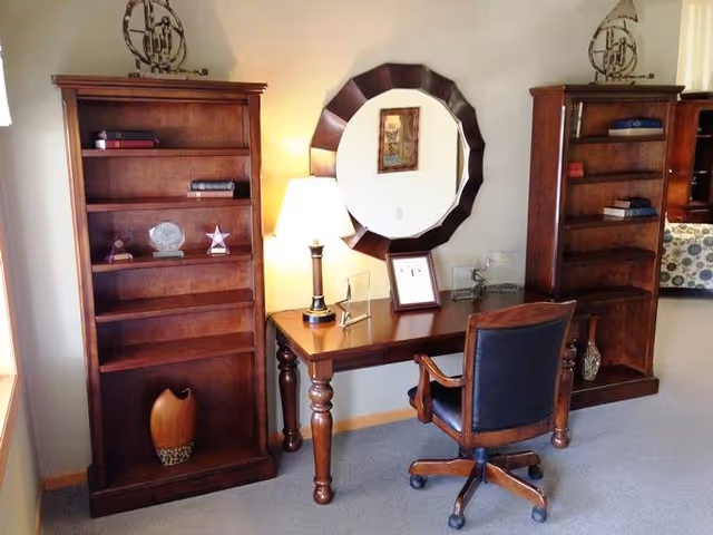 A cozy office or study area featuring a wooden desk with a lamp, framed pictures, and a round decorative mirror on the wall. Two wooden bookshelves flank the desk, each holding a few books and decorative items. A wooden swivel chair with a cushioned seat is positioned in front of the desk. The room has carpeted flooring and neutral-colored walls.