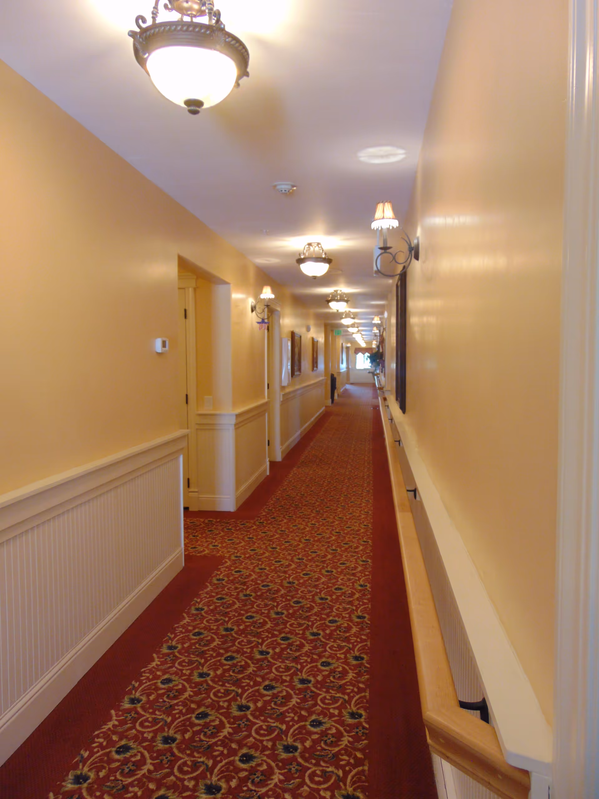 A long, well-lit hallway with beige walls and a red patterned carpet. The hallway features multiple ceiling light fixtures and wall sconces with small lampshades. There are several doorways and framed pictures along the walls, and a wooden handrail runs along the right side.