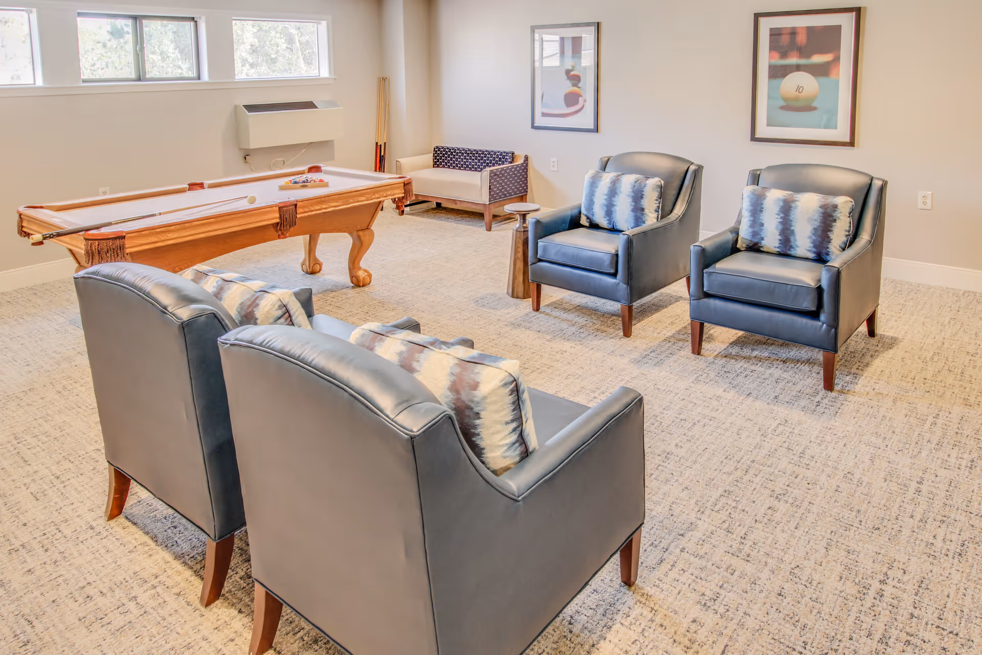 A bright room with a pool table in the center, surrounded by four dark leather armchairs with patterned cushions. There is a small wooden side table between two of the chairs and a small bench against the wall. Two framed pictures of billiard balls hang on the wall, and windows near the ceiling let in natural light.