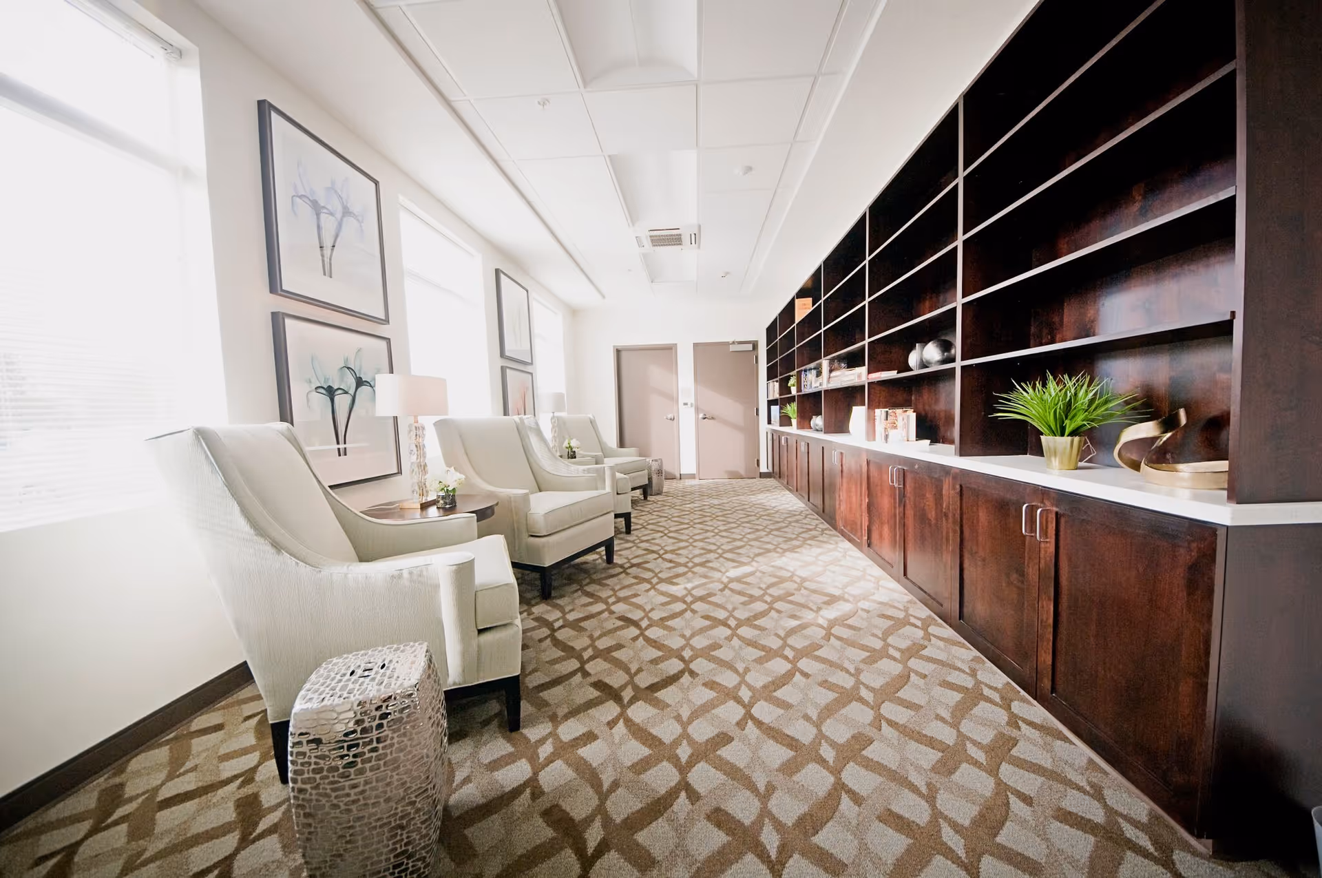 A bright and spacious sitting area with several white upholstered armchairs arranged along a wall with large windows. The opposite wall features dark wooden built-in shelves and cabinets decorated with plants, books, and decorative items. The floor is carpeted with a patterned design, and framed artwork hangs on the wall near the windows.