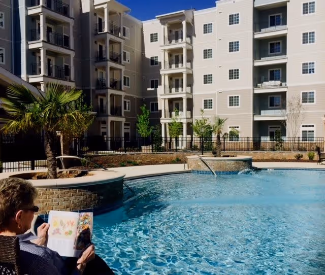 A senior person sitting by a swimming pool reading a book, with a multi-story residential building and palm trees in the background under a clear blue sky.