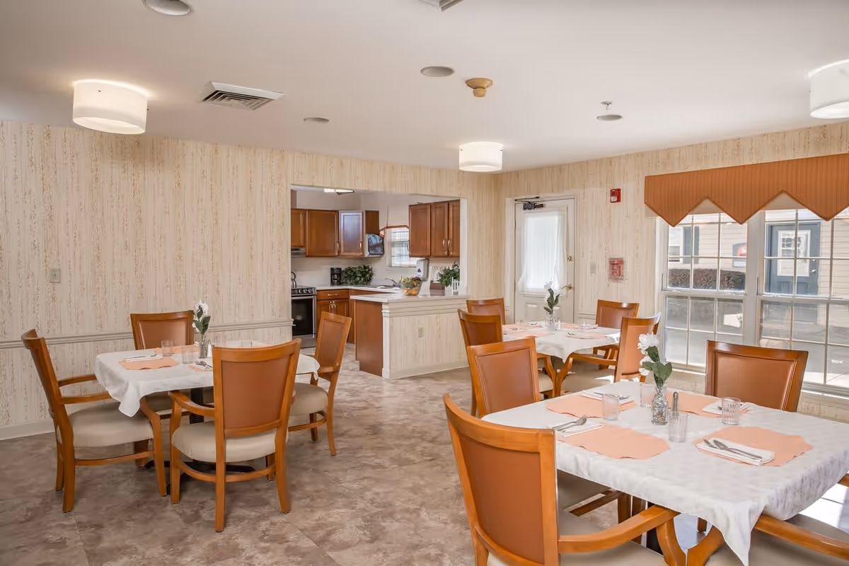 A dining room in a senior living facility with several tables covered with white tablecloths and peach-colored placemats. Each table has wooden chairs with cushioned seats and a small vase with flowers. The room has beige patterned wallpaper, large windows with a brown valance, and ceiling lights. In the background, there is an open kitchen area with wooden cabinets and appliances.