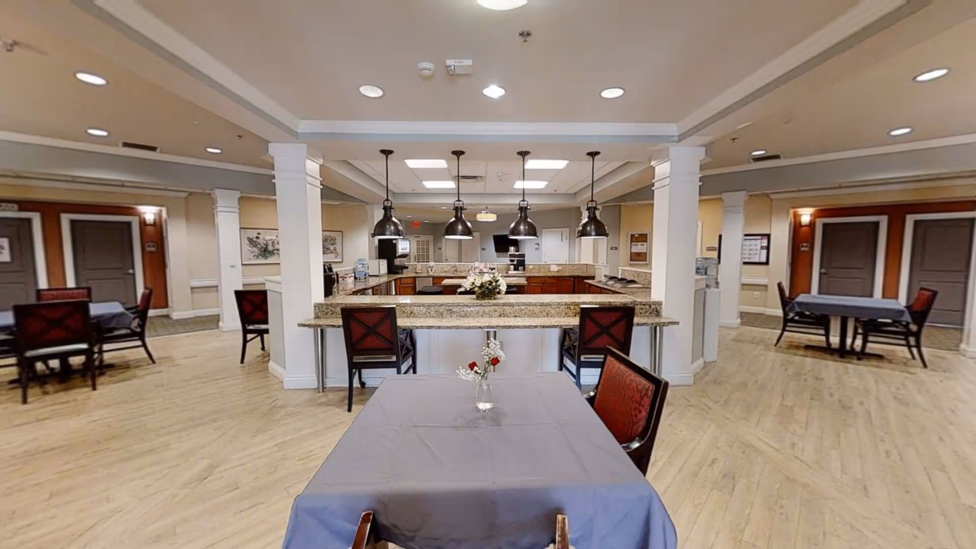 Interior view of a senior living facility dining area with tables covered in gray tablecloths and chairs with red cushions. In the center is a kitchen island with granite countertops and four hanging pendant lights above. The room has light wood flooring, white columns, and beige walls with doors and framed artwork in the background.