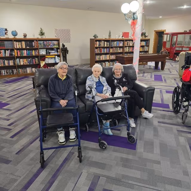 Three elderly women sitting on a black leather couch in a spacious room with patterned carpet. Two of the women have walkers in front of them. Behind them are bookshelves filled with books and some decorative items. A pool table and a lamp post with round lights are visible in the background.