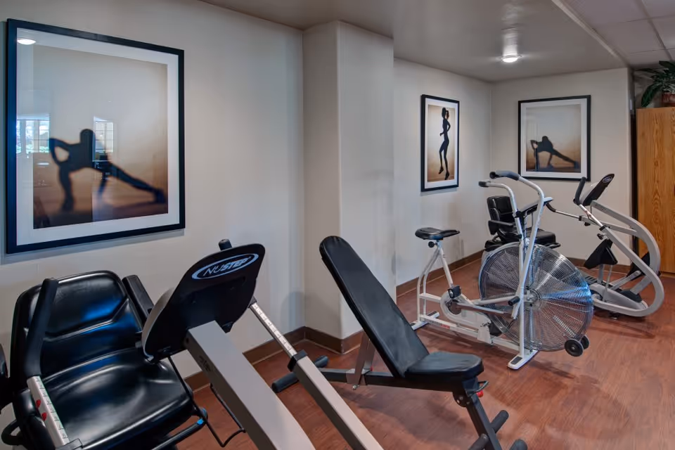 Interior view of a fitness room with exercise equipment including a recumbent bike, a stationary bike, and an elliptical machine. The room has wooden flooring and white walls adorned with framed silhouette artwork of people exercising.