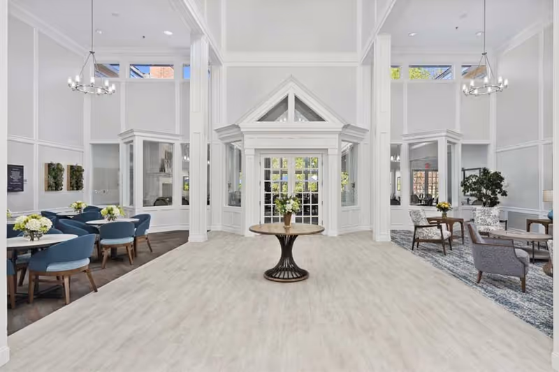 Bright and spacious common area with high ceilings and large windows. The room features a central round table with a flower arrangement, multiple seating areas with blue and patterned chairs, and small tables with flower decorations. White walls and light wood flooring create an airy atmosphere.