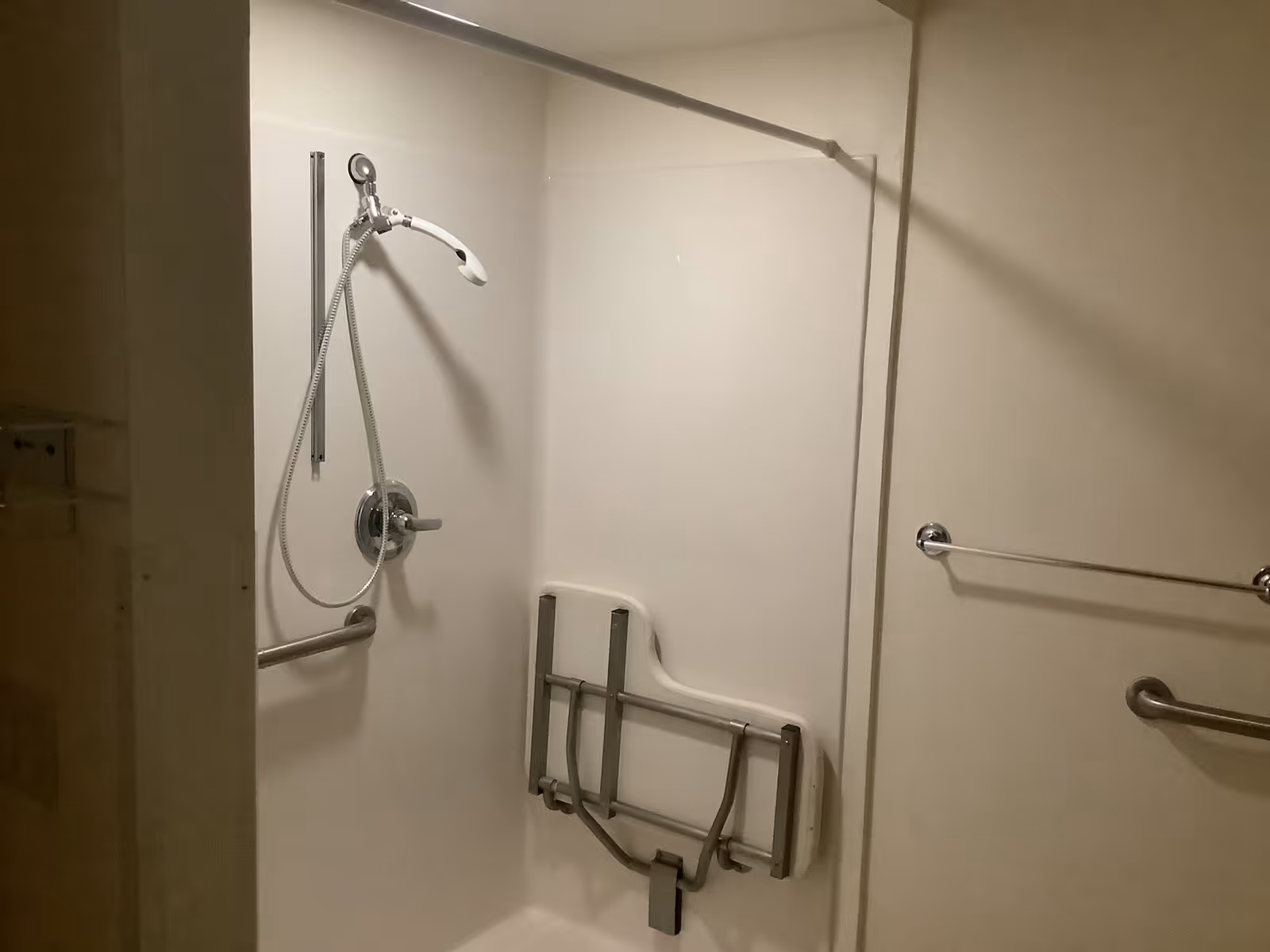 A shower area with a handheld showerhead attached to a sliding bar, a foldable shower seat mounted on the wall, and two grab bars for support. The walls and fixtures are white and chrome, designed for accessibility.