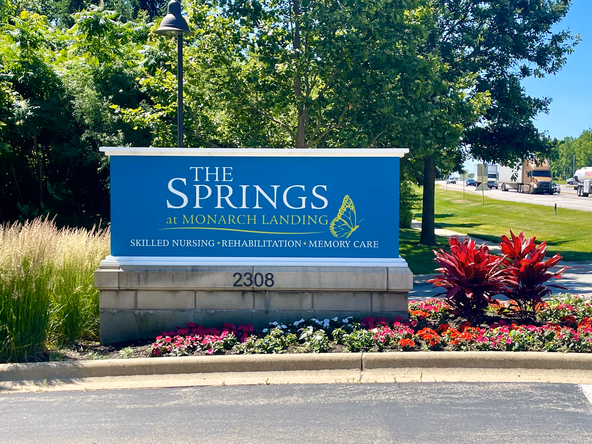 Outdoor sign for The Springs at Monarch Landing, a skilled nursing, rehabilitation, and memory care facility, surrounded by greenery and colorful flowers with a road and vehicles in the background.