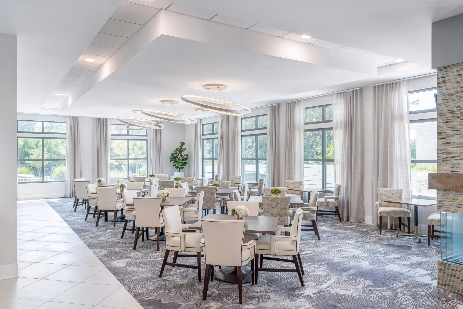 Bright and spacious dining room with multiple tables and chairs arranged neatly. Large windows with sheer curtains allow natural light to fill the room. Modern circular light fixtures hang from the ceiling, and small plants decorate the tables. The room has a patterned carpet and a tiled walkway on the left side.
