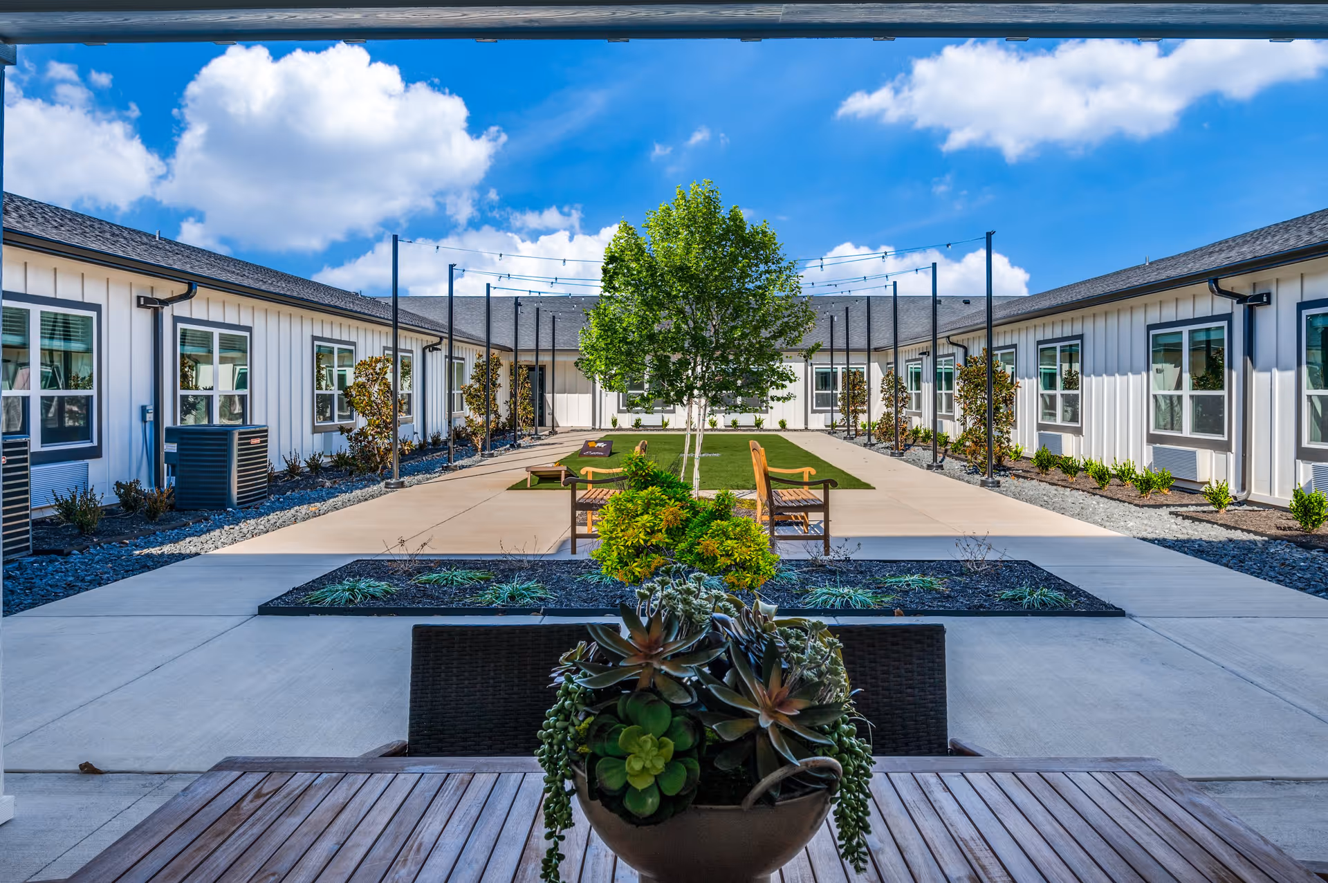 Central courtyard of a senior living facility with a paved walkway, benches, planted beds, a central tree, and surrounding single-story buildings under a blue sky.
