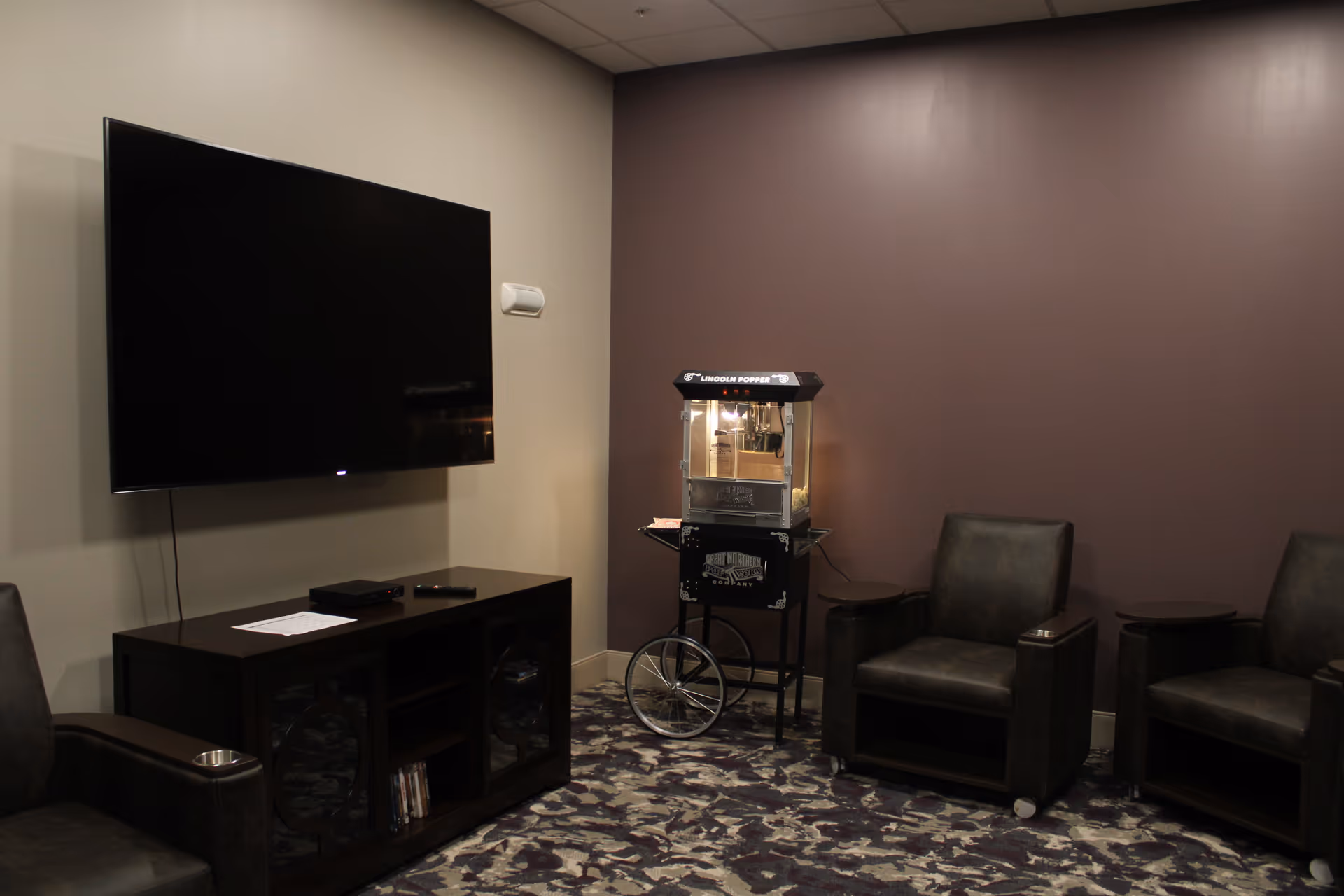 A cozy lounge area with a large flat-screen TV mounted on a beige wall, a dark wooden TV stand below it, a vintage-style popcorn machine on wheels, and three dark leather armchairs arranged around the popcorn machine. The walls are painted beige and dark brown, and the floor is covered with a patterned carpet.