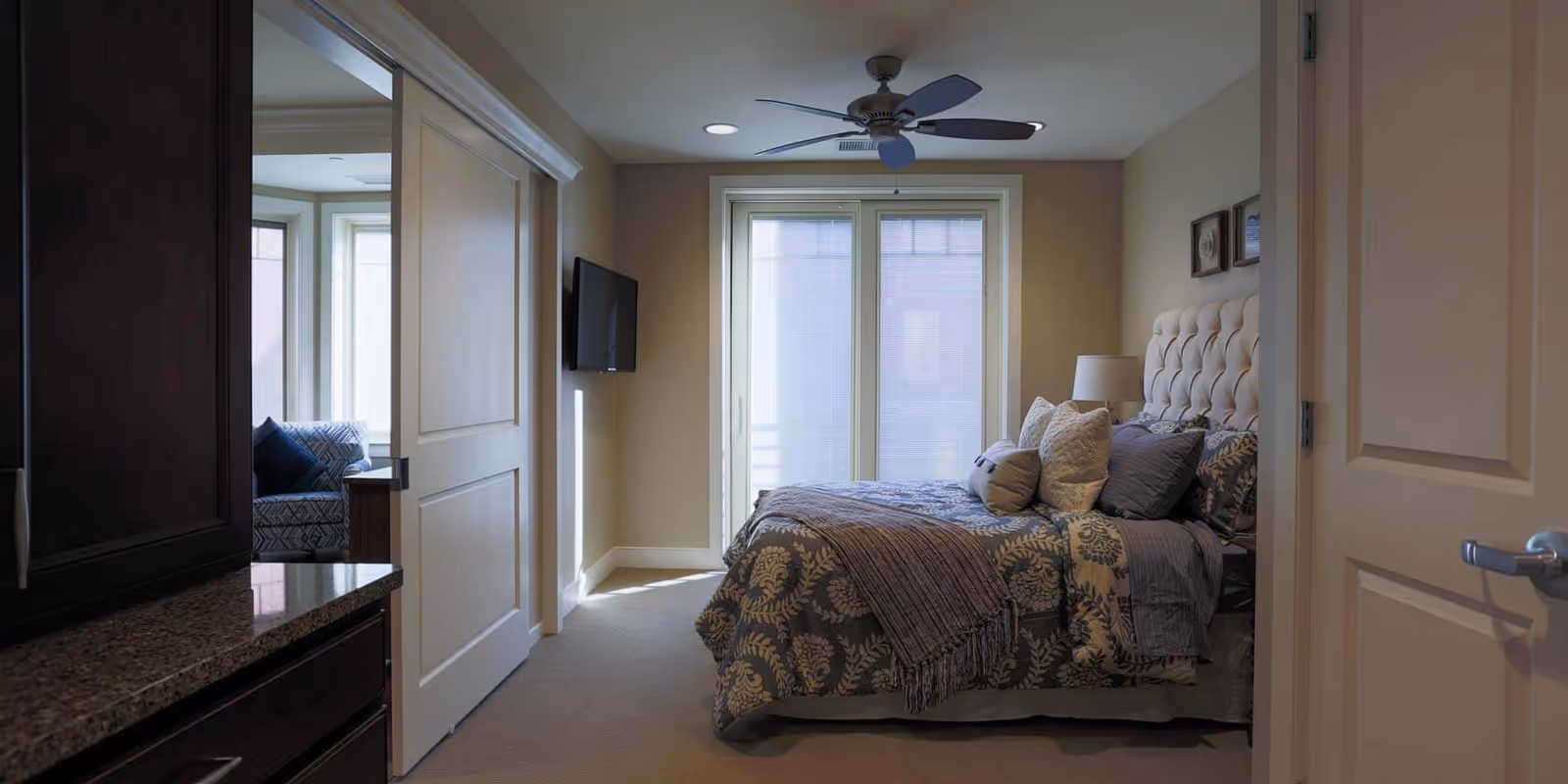 Cozy bedroom with a tufted-headboard bed dressed in patterned bedding, a wall-mounted TV, ceiling fan, and sliding glass doors letting in light.